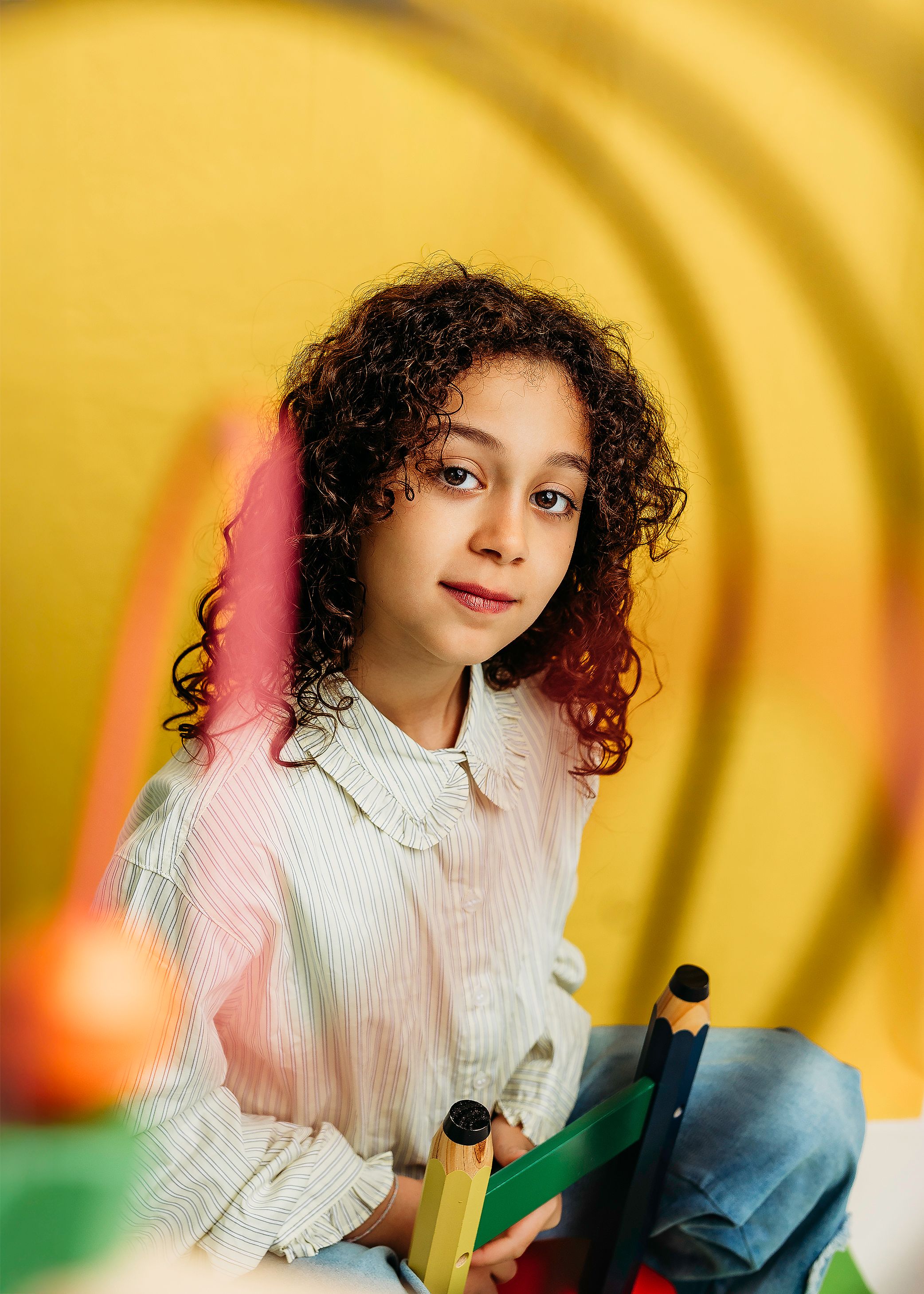 Young girl with curly hair holding oversized pencils against a vibrant yellow background.