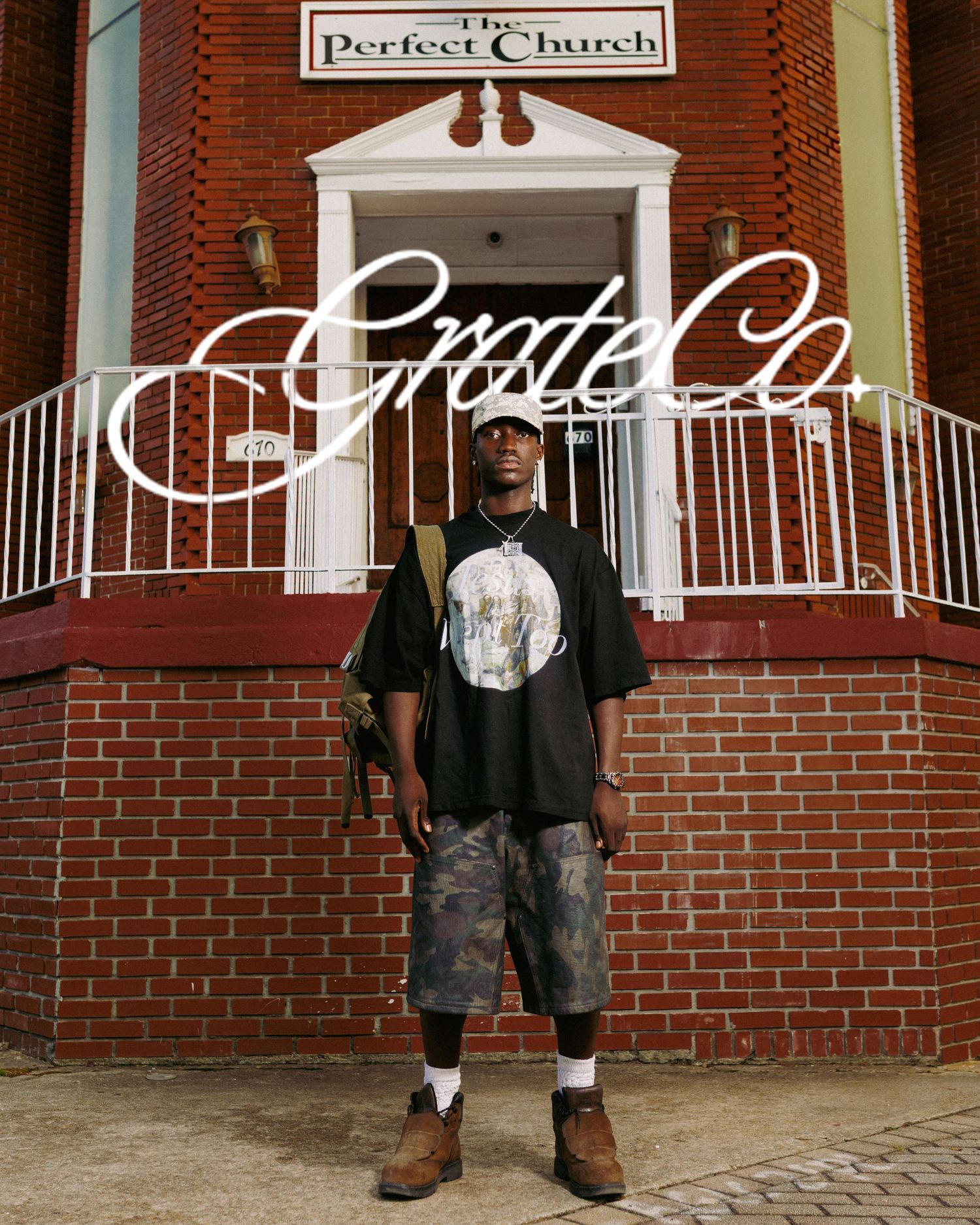 A young man stands confidently in front of The Perfect Church brick building wearing a black graphic tee and camo shorts.