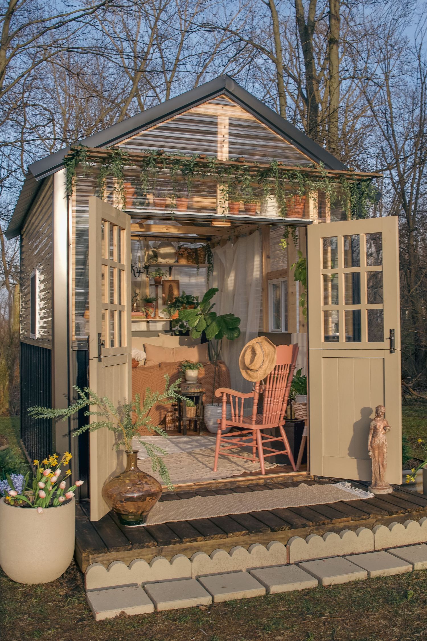 Charming garden shed with open French doors, pink rocking chair, lush plants, and cozy interior, surrounded by bare trees in soft sunlight.