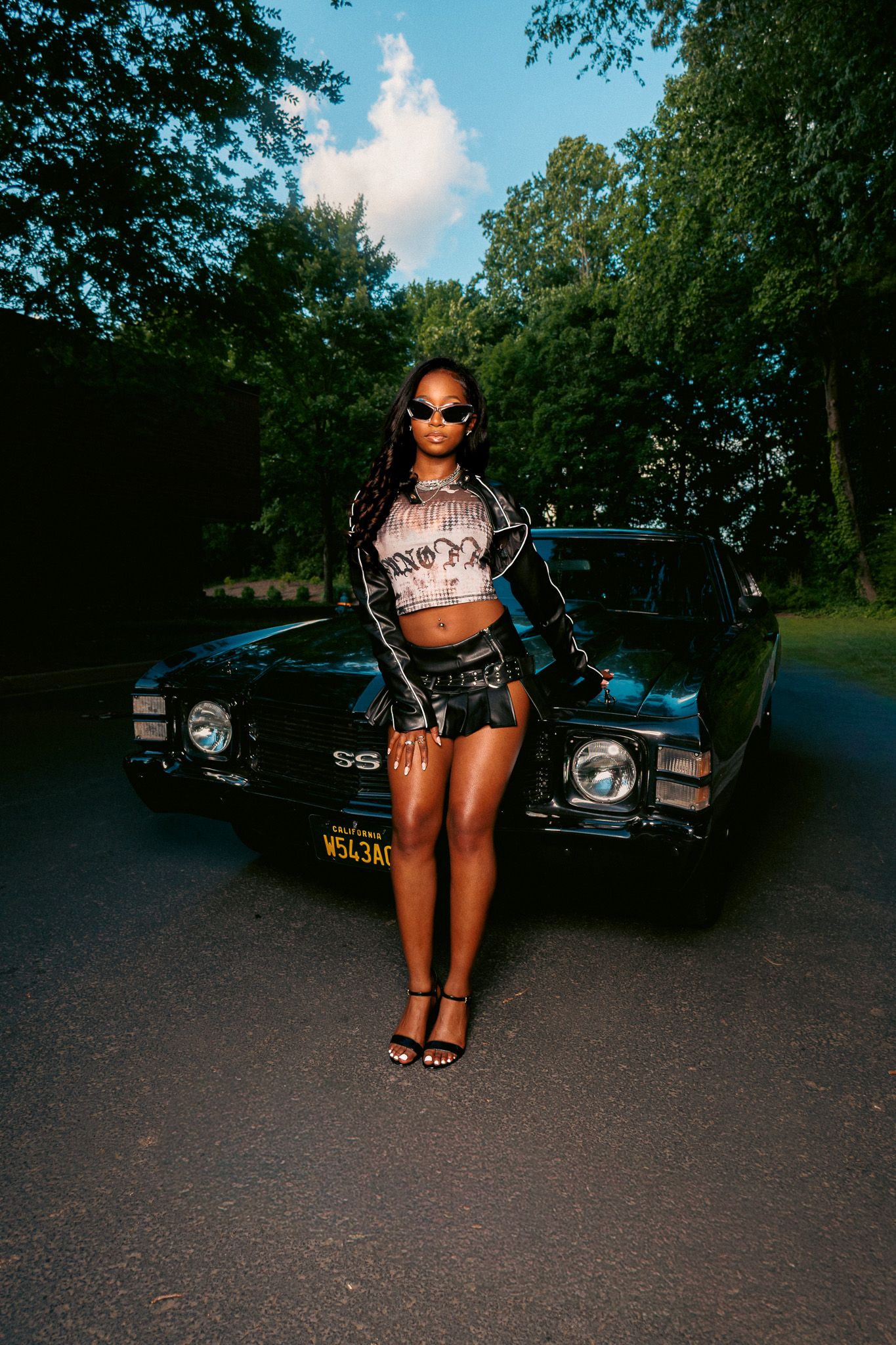 A stylish woman in leather shorts and crop top poses by a classic black SS muscle car on a tree-lined road.