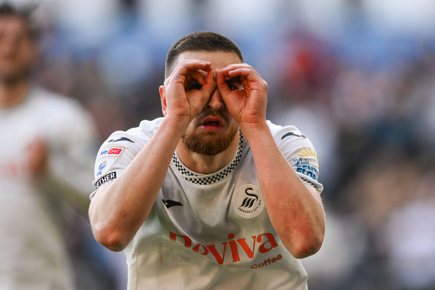A soccer player in a white Swansea City jersey makes binoculars gesture with his hands.
