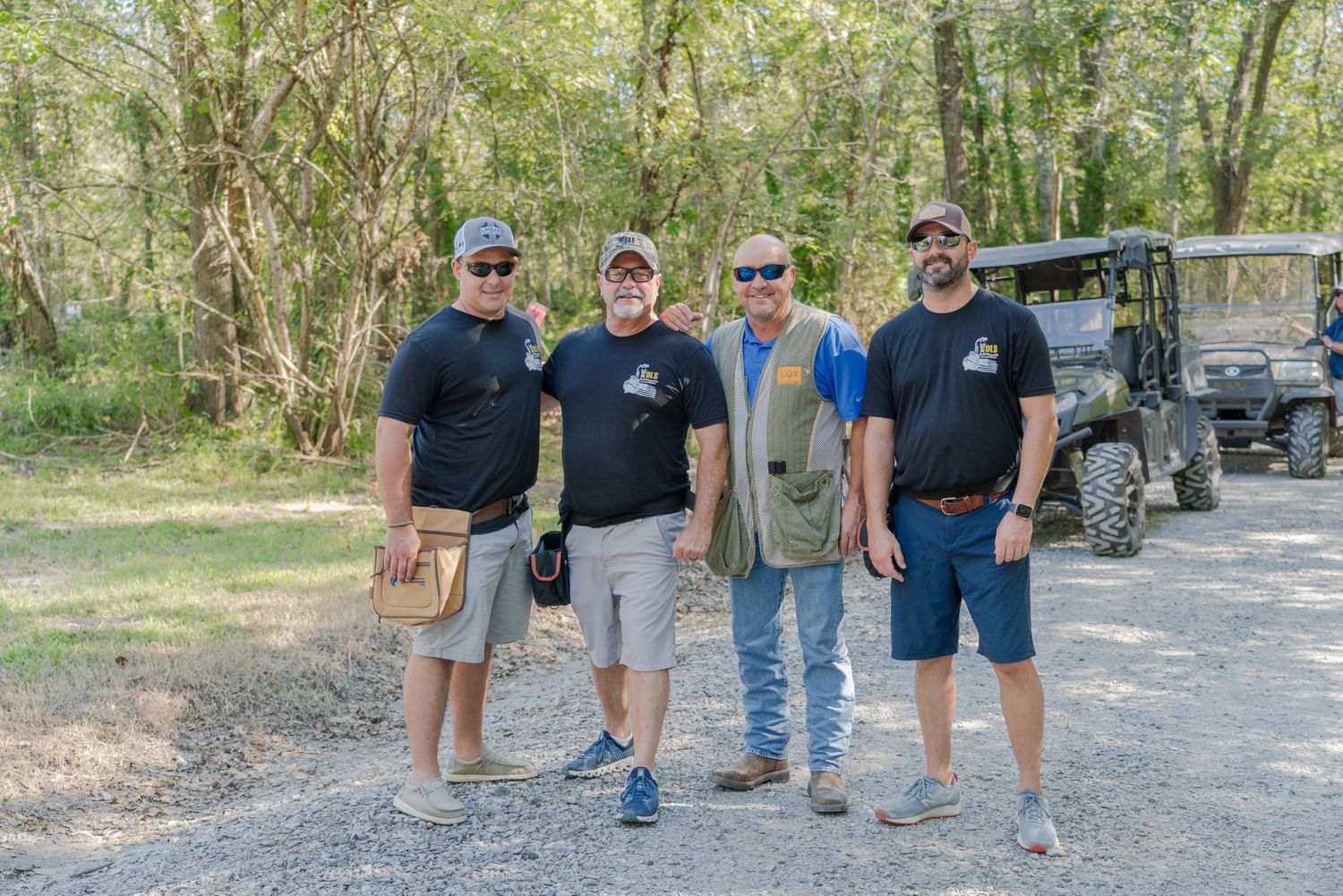 Four men in matching black shirts pose together on a gravel path surrounded by trees near an ATV.