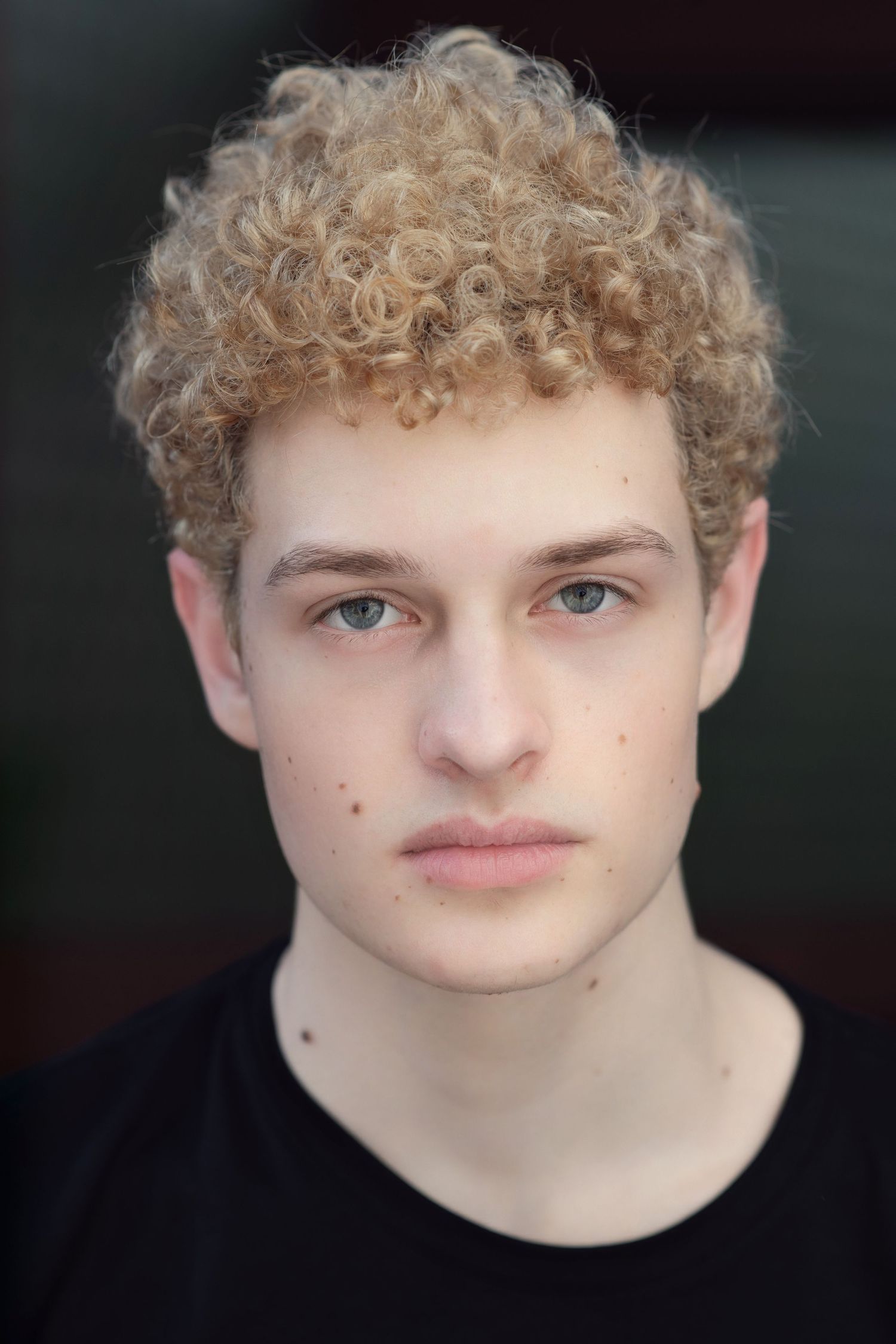 Young man with curly blonde hair and blue eyes wearing a black t-shirt against a dark background.