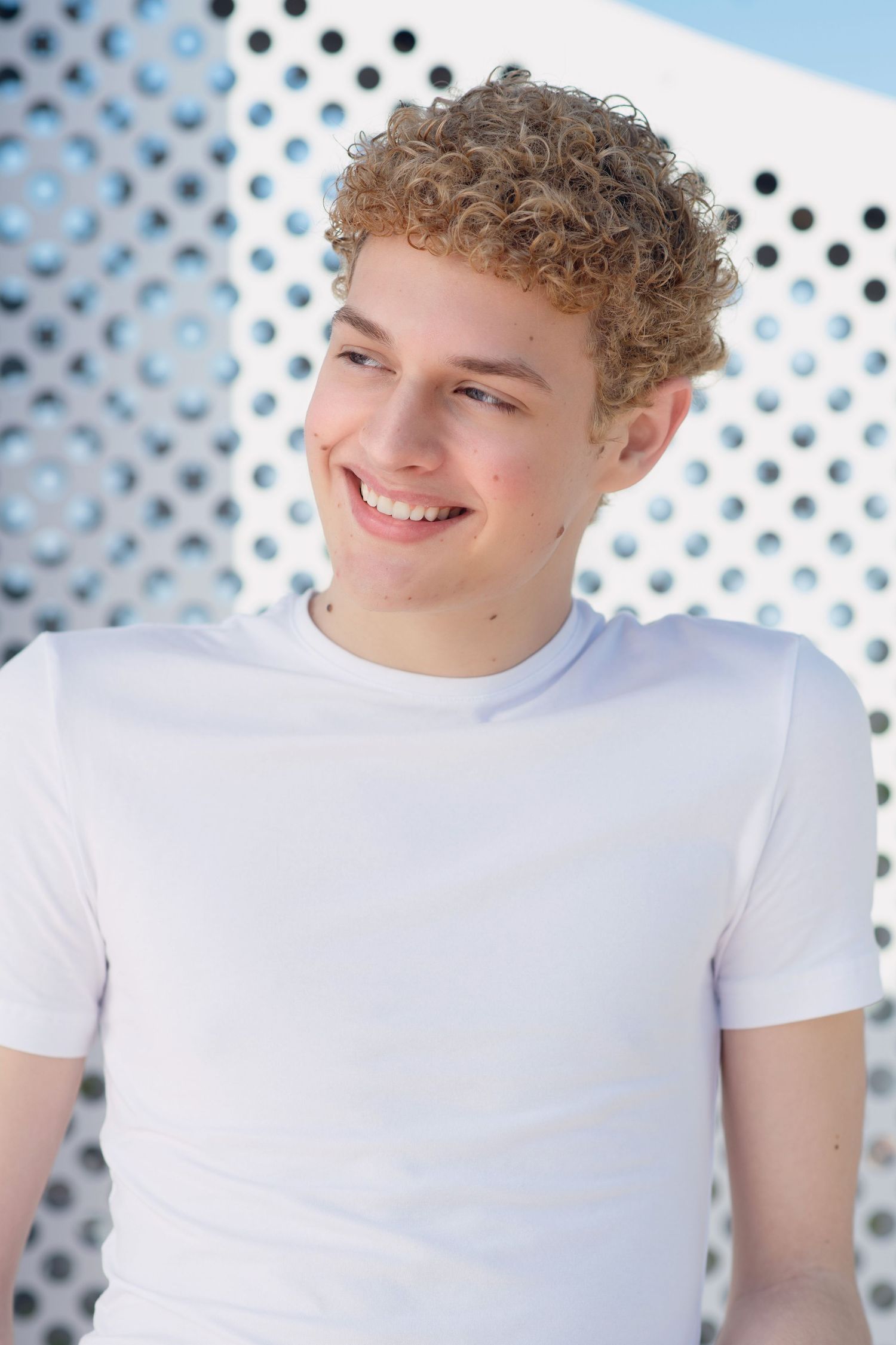 Smiling young man with curly blonde hair wearing a white t-shirt against latticed wall.