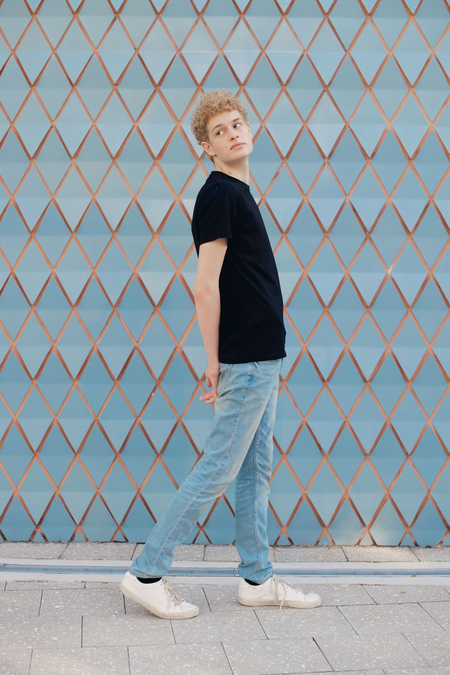 Young man with curly blonde hair walking in black tee and light jeans against blue diamond-patterned wall.