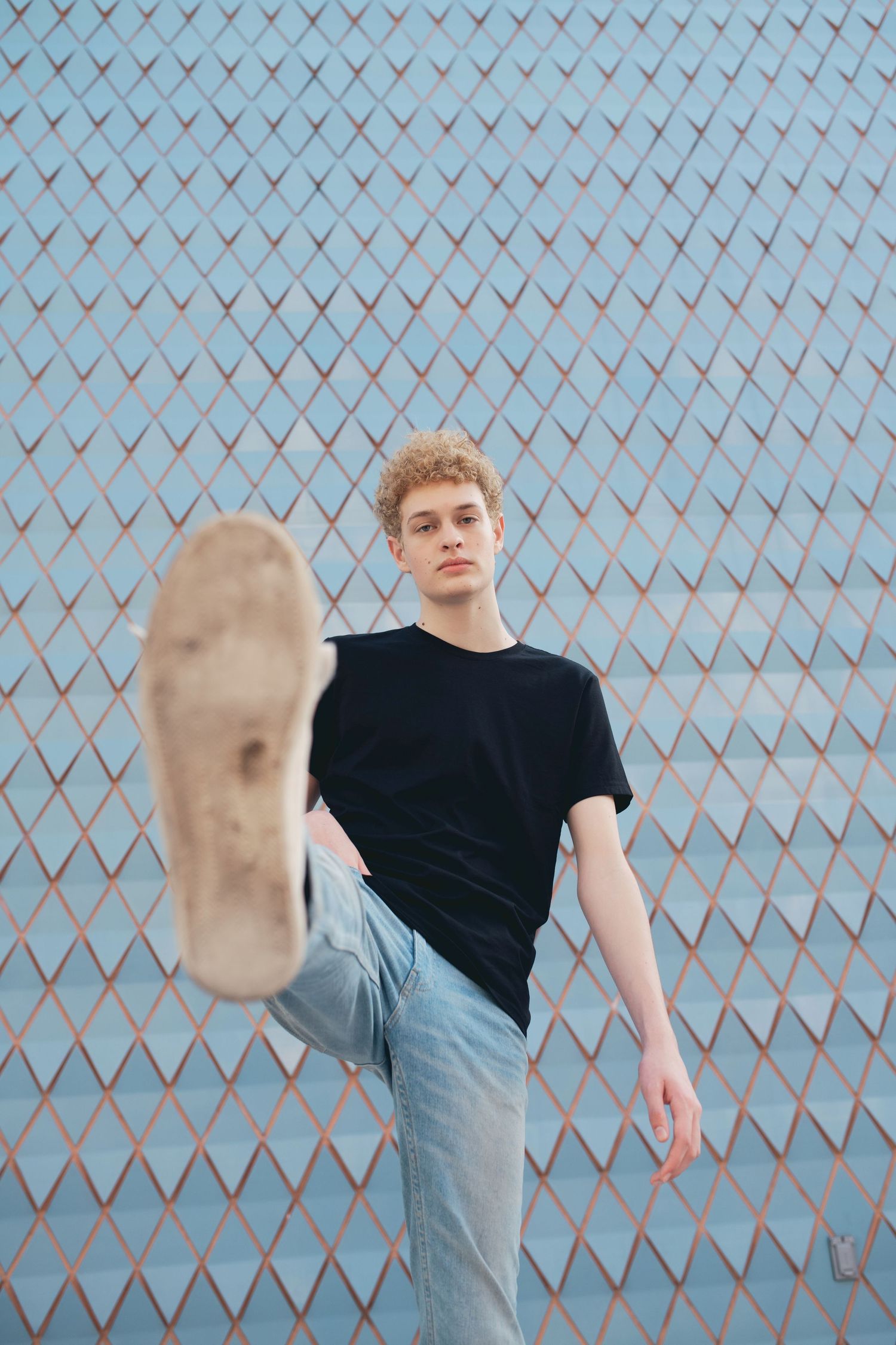 Young curly-haired male model kicking leg toward camera in black t-shirt and jeans against blue grid wall.