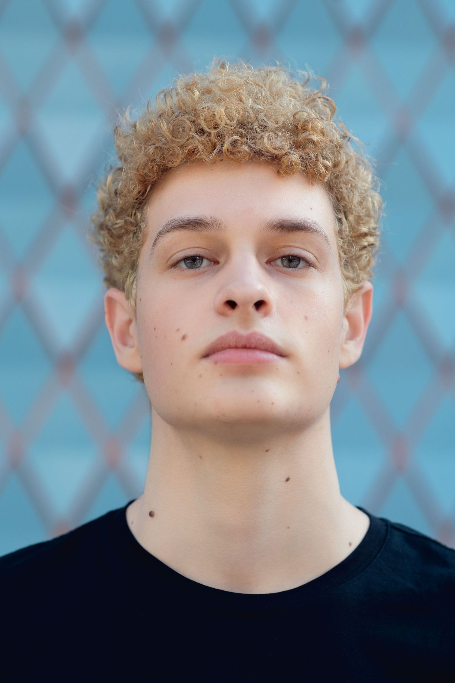 Close-up portrait of young man with curly blonde hair and freckles wearing a black t-shirt outdoors.
