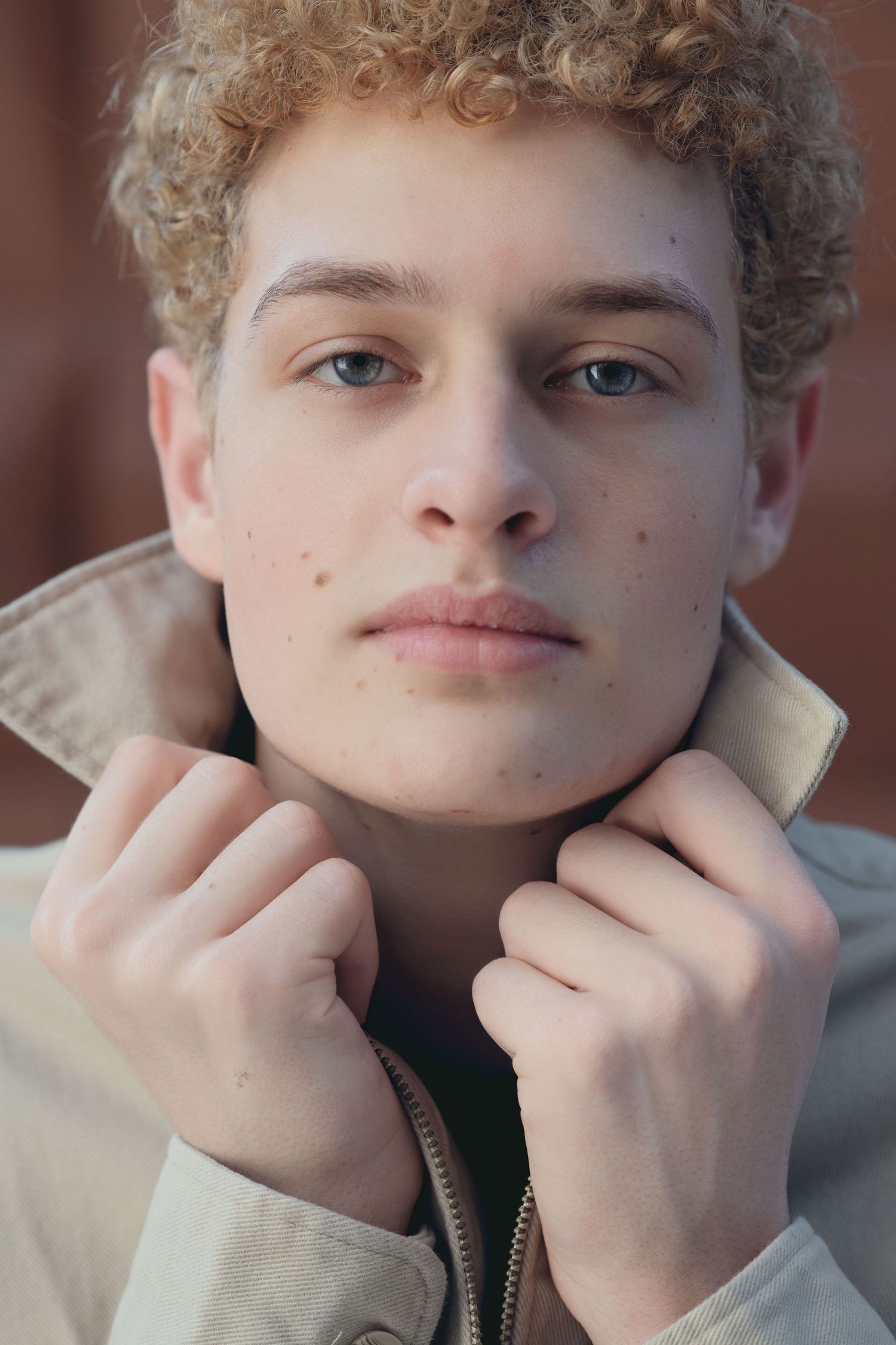 Close-up portrait of curly-haired young man adjusting beige jacket collar with both hands.