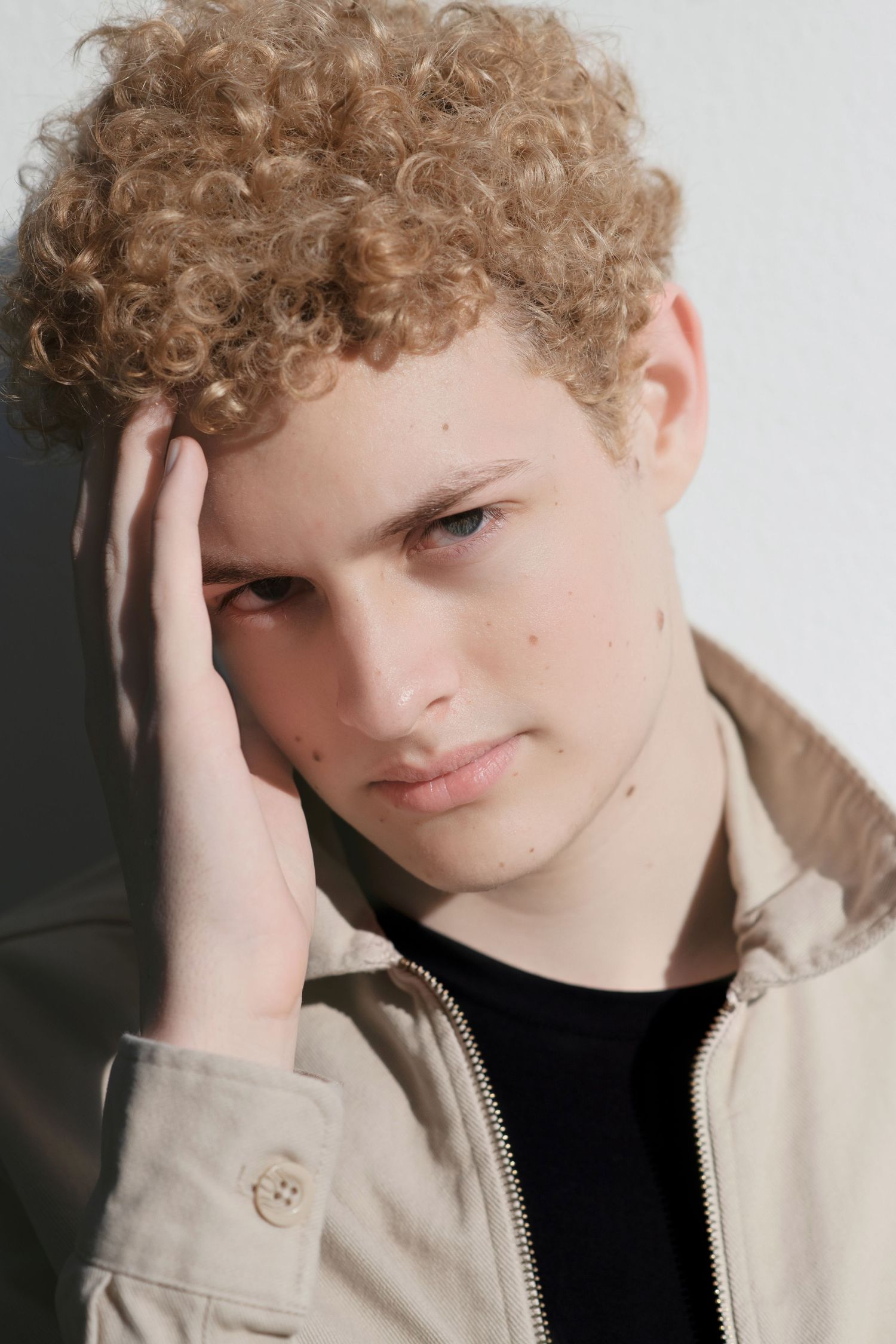Pensive young man with curly blonde hair resting hand on head wearing beige zip jacket indoors.