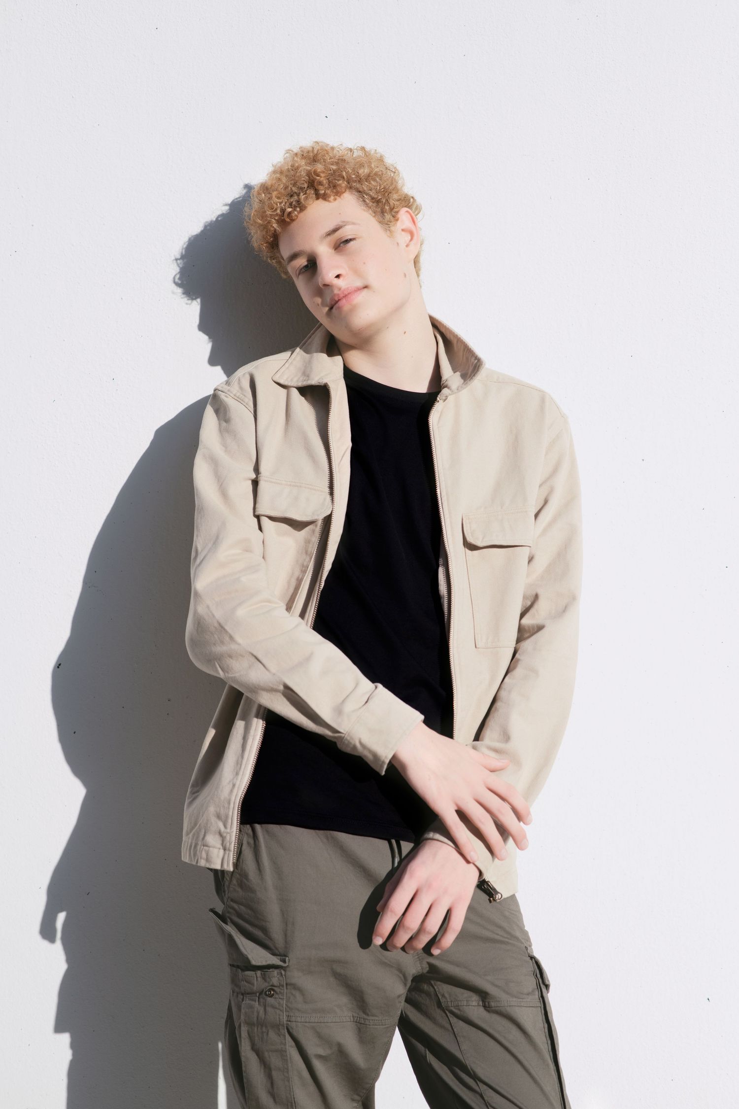 Young man with curly blonde hair leaning against white wall in beige jacket and olive cargo pants.