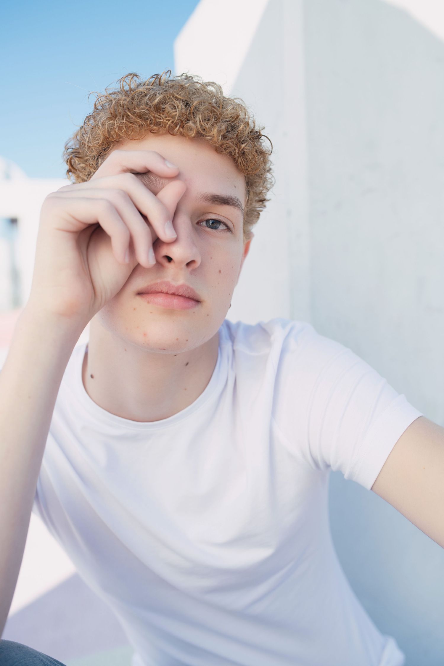 Young man with curly blonde hair in white t-shirt posing thoughtfully outdoors.