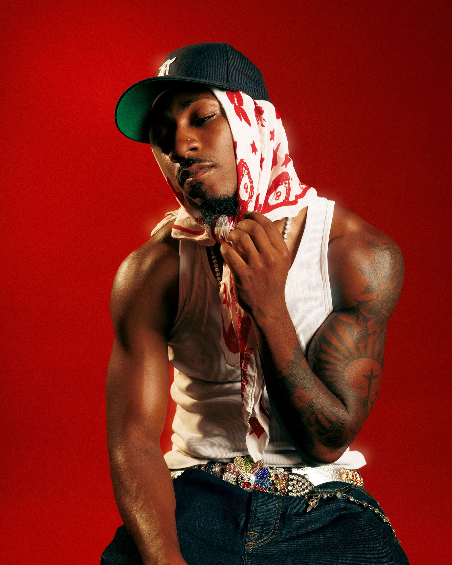 Muscular man in white tank top, blue cap, red bandana, and jeans poses against a red background with tattoos visible.