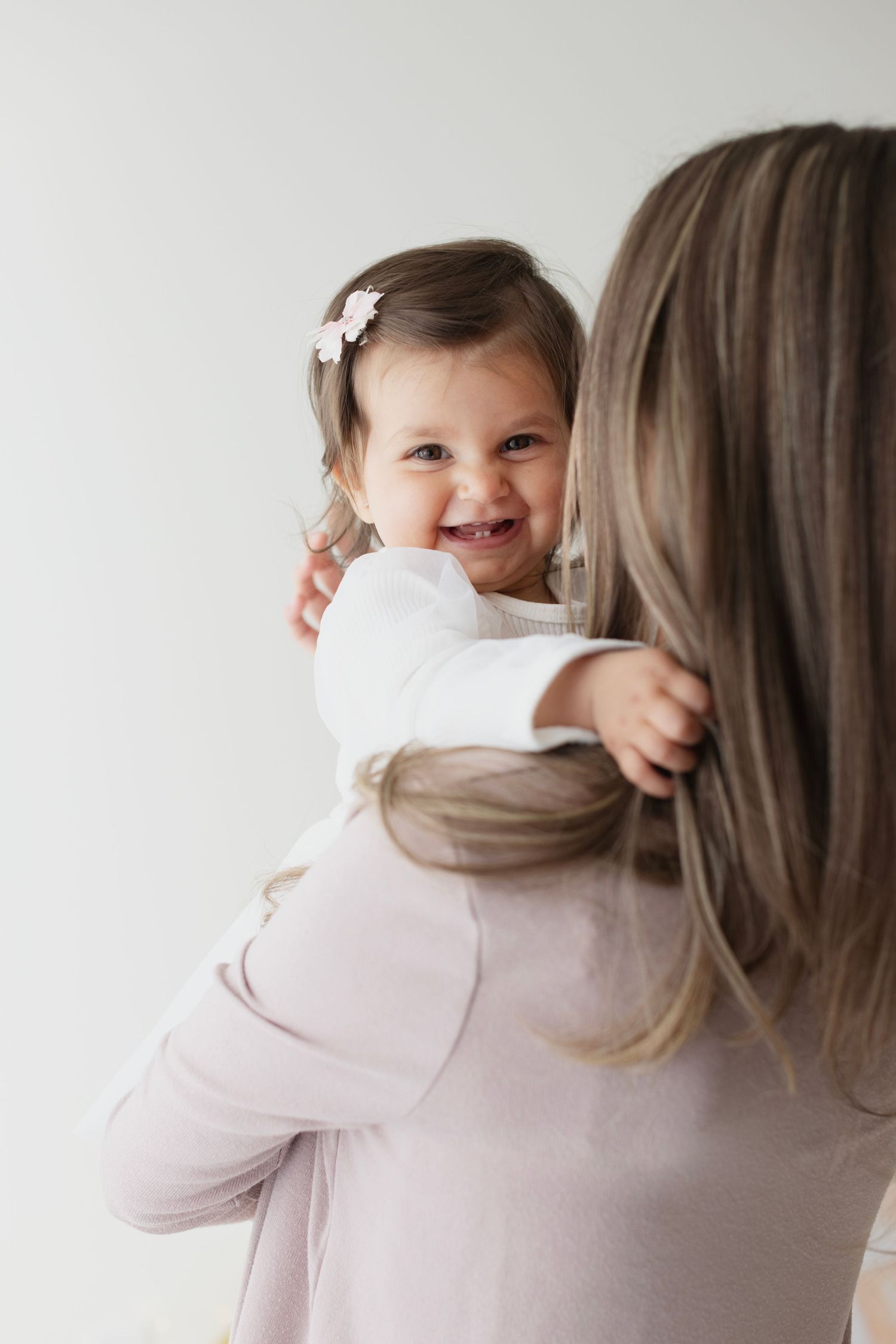 A smiling toddler girl with a pink hair clip is held over the shoulder of a woman with long blonde hair. Elizabeth Lang Photography Surrey