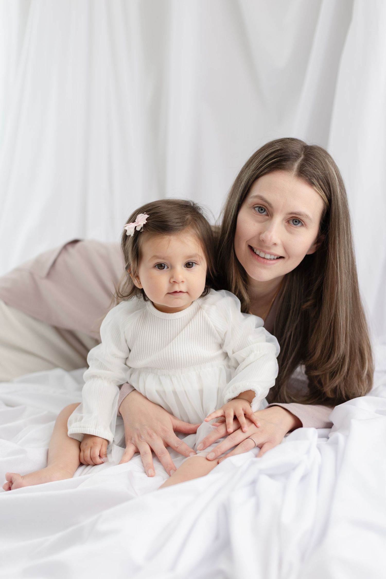 A smiling mother lies on white bedding while gently holding her baby girl in a white dress with a flower hair clip. Elizabeth Lang Photograp