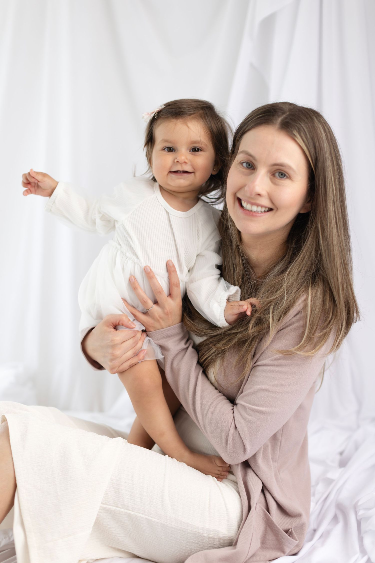 Smiling mother holding happy toddler in white outfit against white background. Elizabeth Lang Photography Surrey