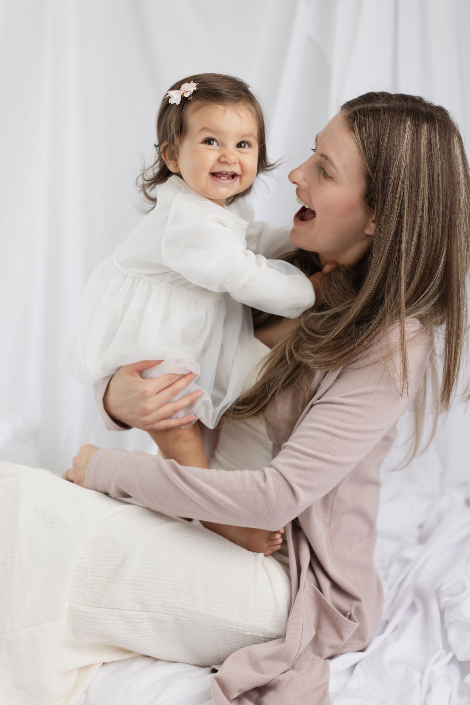 A joyful mother in blush cardigan playfully holding her smiling baby girl in a white dress against soft white backdrop. Elizabeth Lang Photo