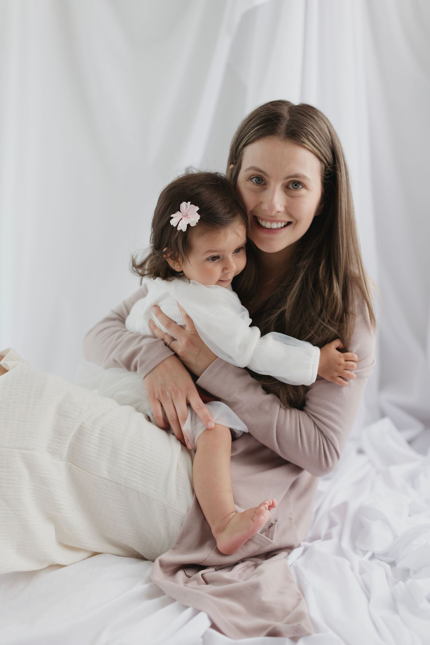 A happy mother in a pink dress sits on white bedding, warmly embracing her toddler daughter with a floral hair clip. Elizabeth Lang Photogra