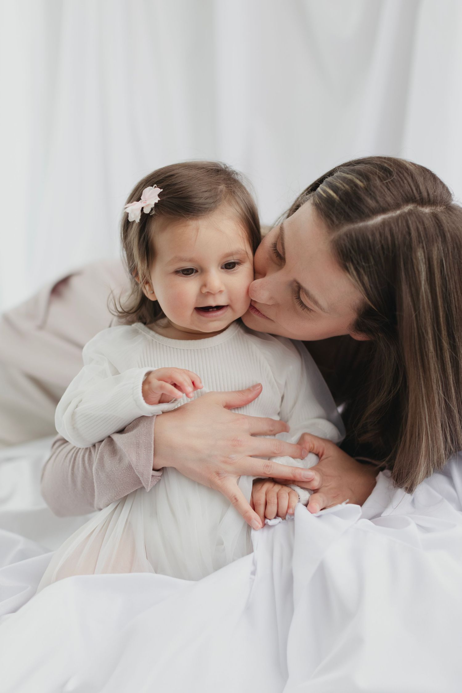 A loving mother kisses her smiling baby girl in a white dress on the cheek while sitting on white bedding. Elizabeth Lang Photography Surrey