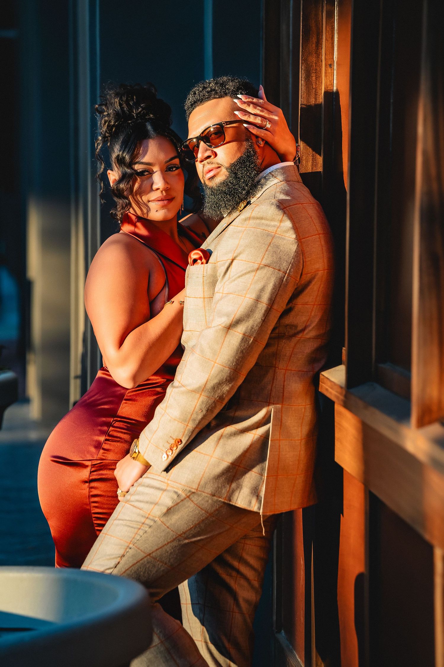 Stylish couple in warm lighting pose together against a wall, with her in red and him in beige formal wear.