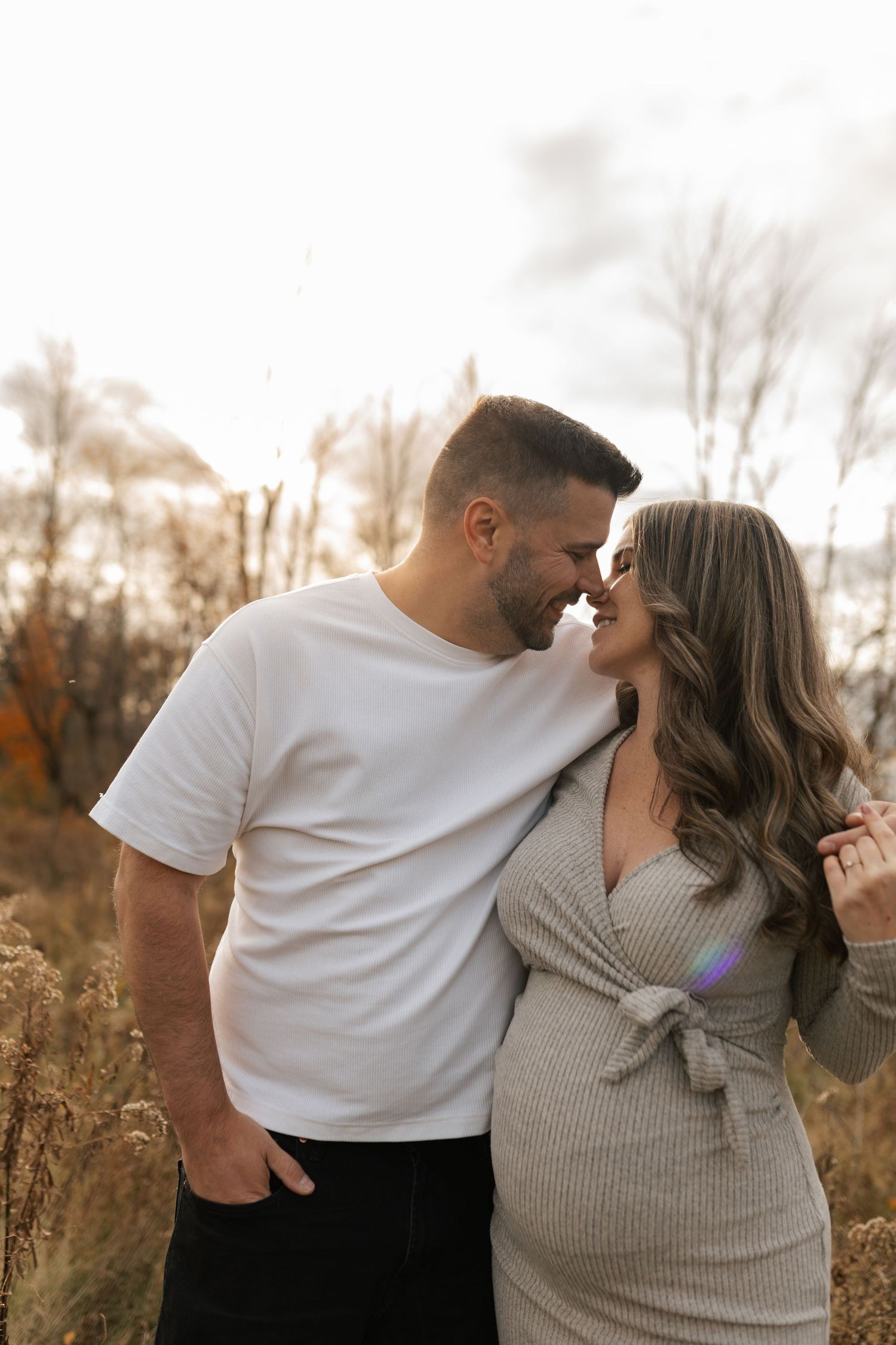 Expecting couple sharing tender nose-to-nose moment in golden autumn field with sunlight behind them.