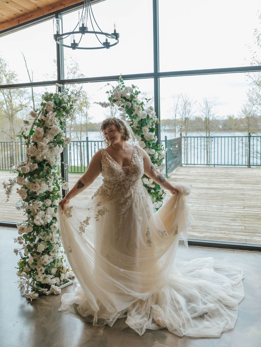 Bride in a floral lace tulle wedding gown poses by a white flower arch with large windows overlooking a lake.
