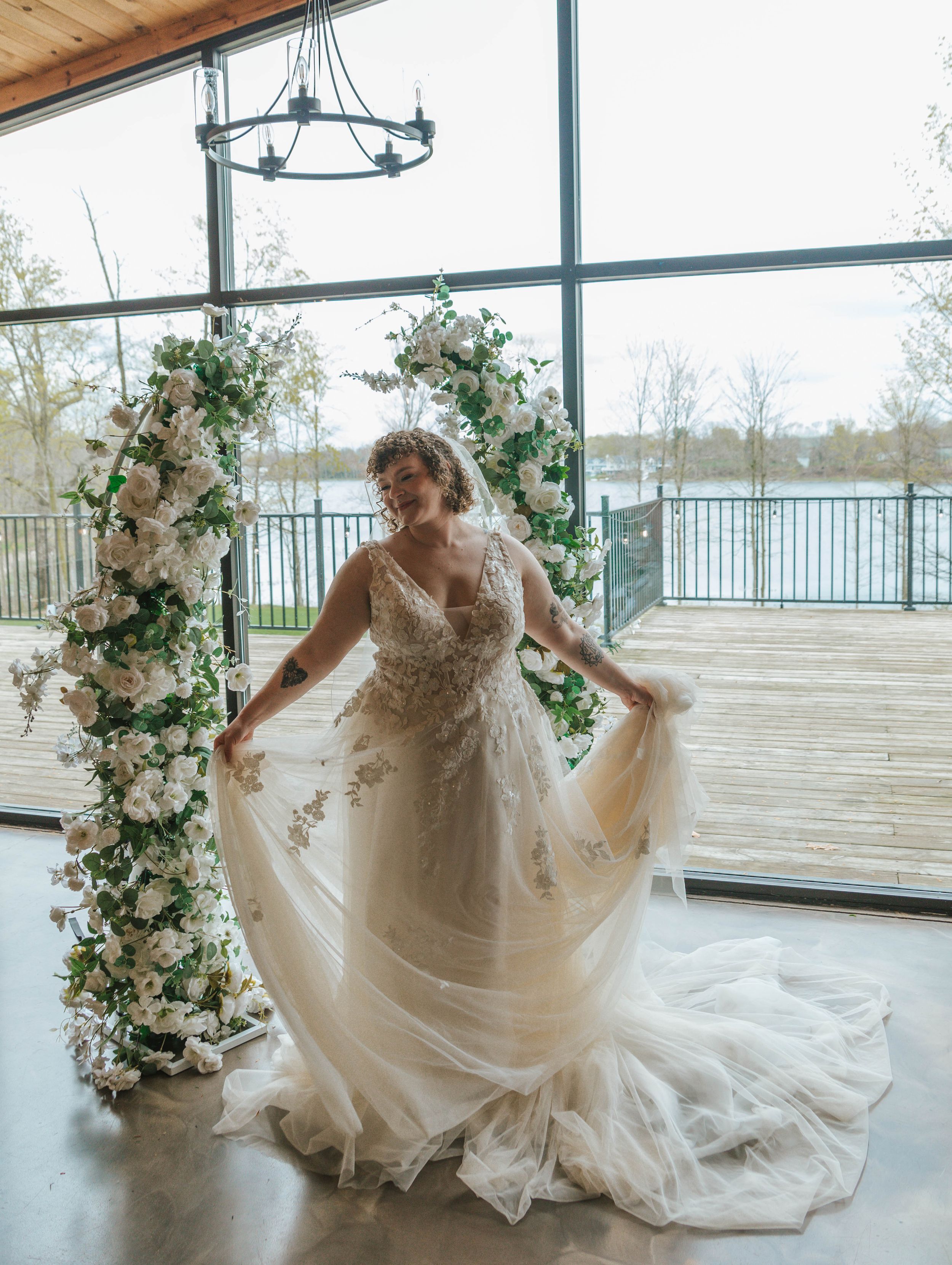 Bride in a floral lace tulle wedding gown poses by a white flower arch with large windows overlooking a lake.