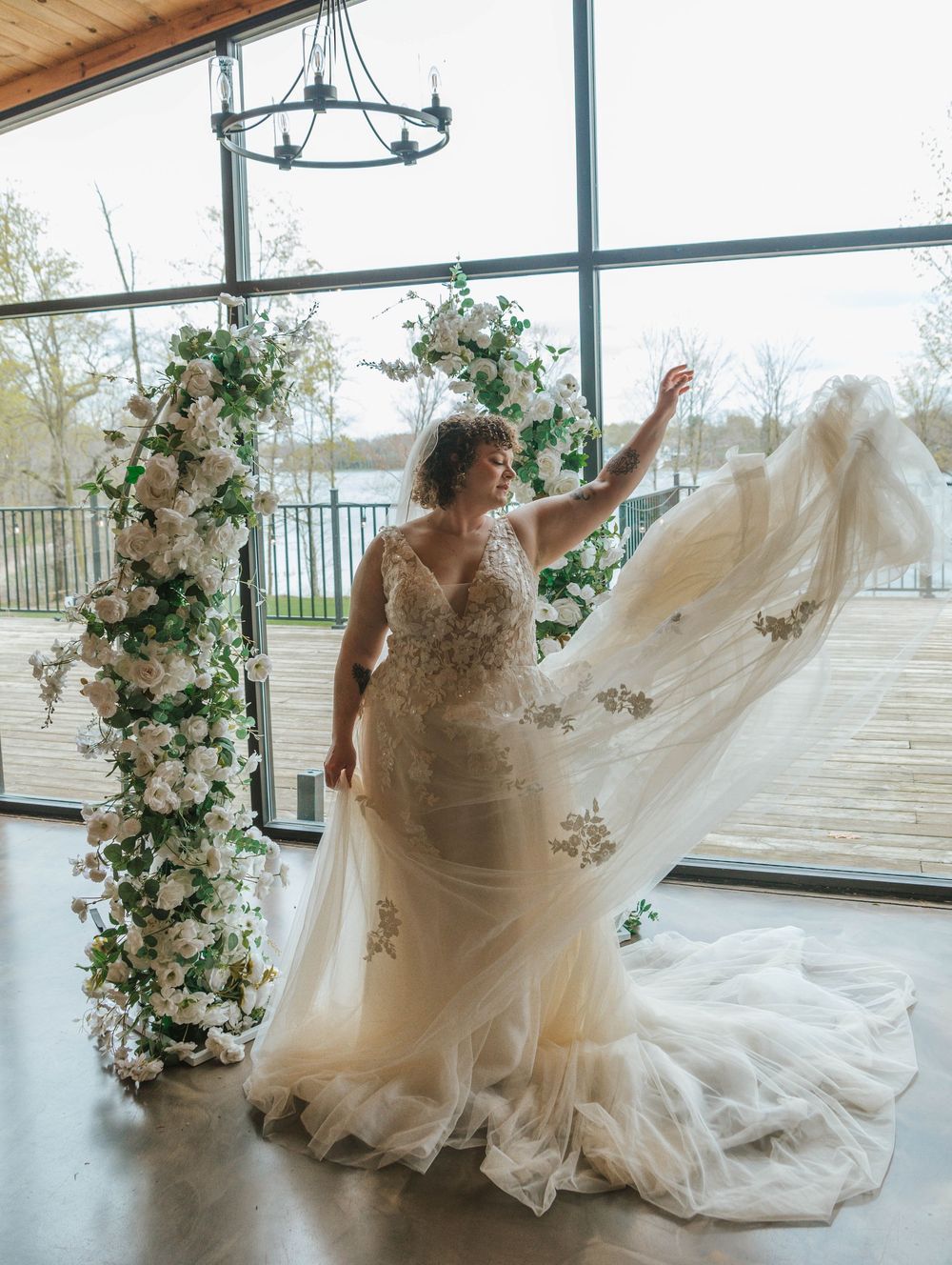 Bride in lace wedding gown with flowing tulle train poses before floral arch columns in sunlit venue.