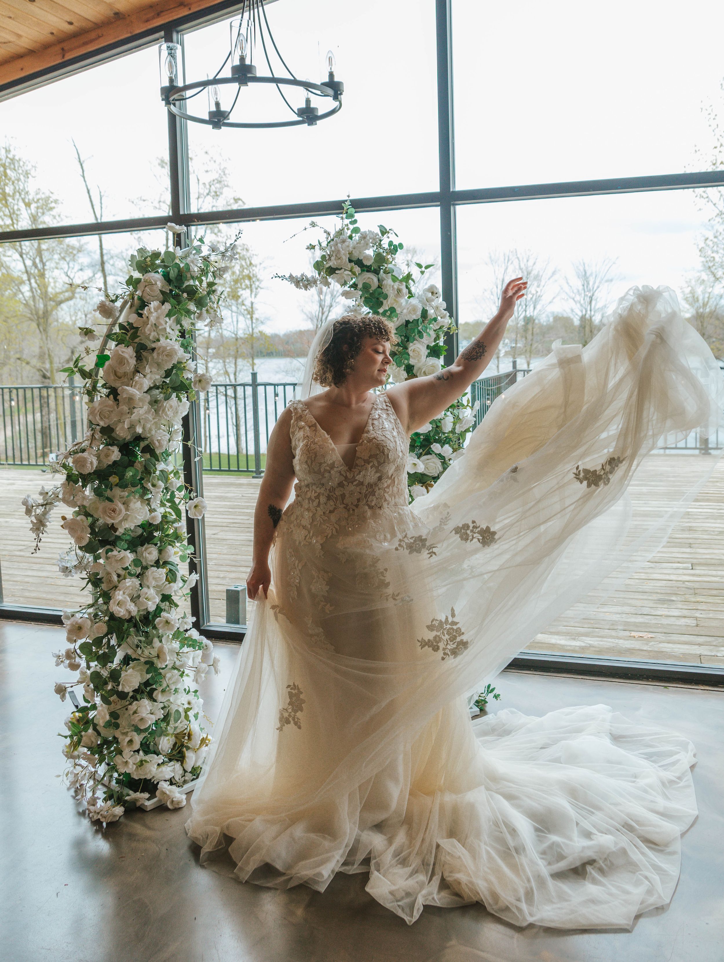 Bride in lace wedding gown with flowing tulle train poses before floral arch columns in sunlit venue.
