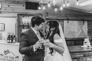 Black and white photo of newlyweds toasting with champagne flutes, nearly kissing at their rustic barn wedding reception.