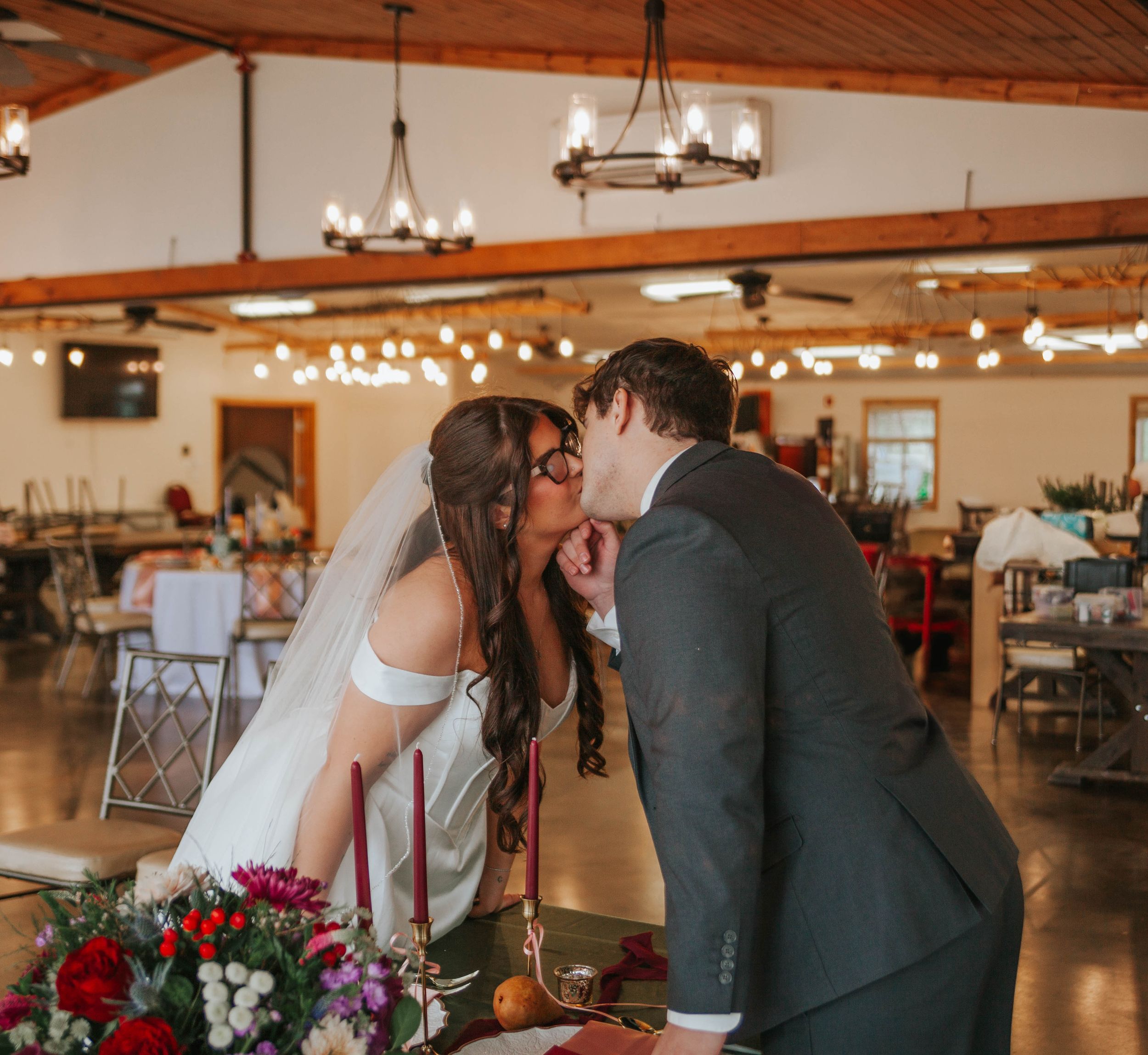 Bride and groom sharing a kiss at wedding reception in rustic barn venue with string lights and floral centerpieces.