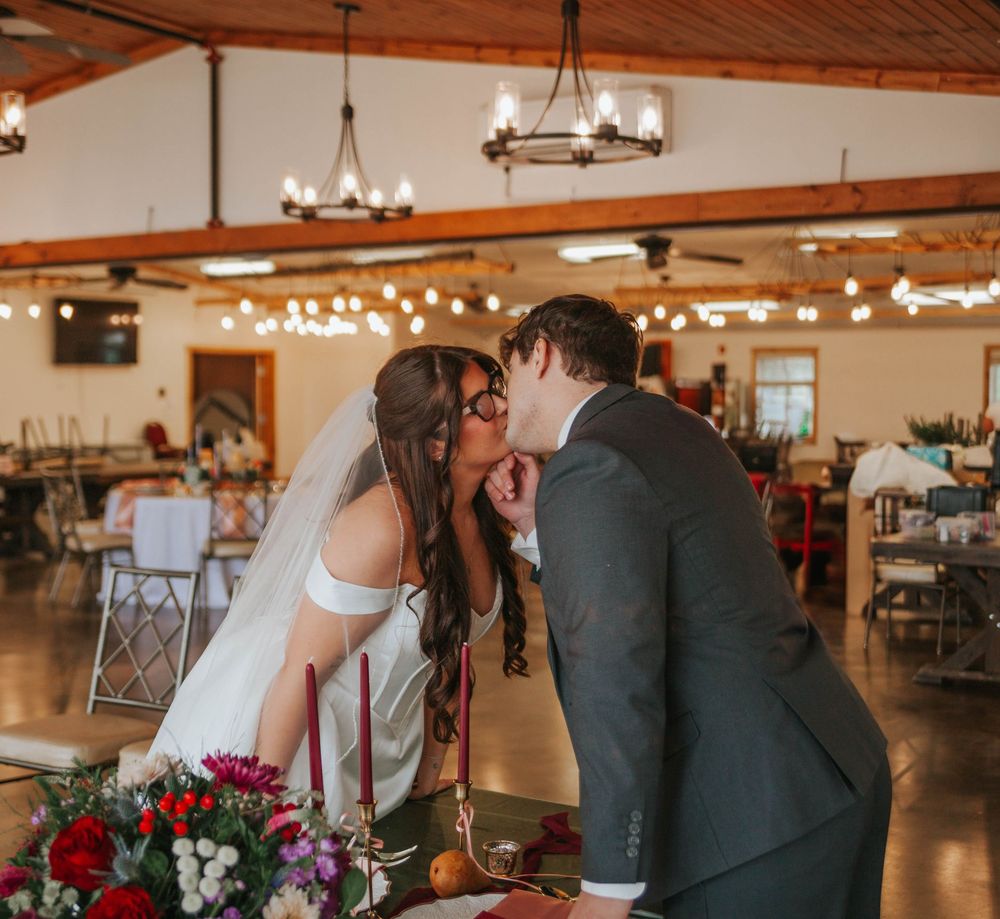 Bride and groom sharing a kiss at wedding reception in rustic barn venue with string lights and floral centerpieces.