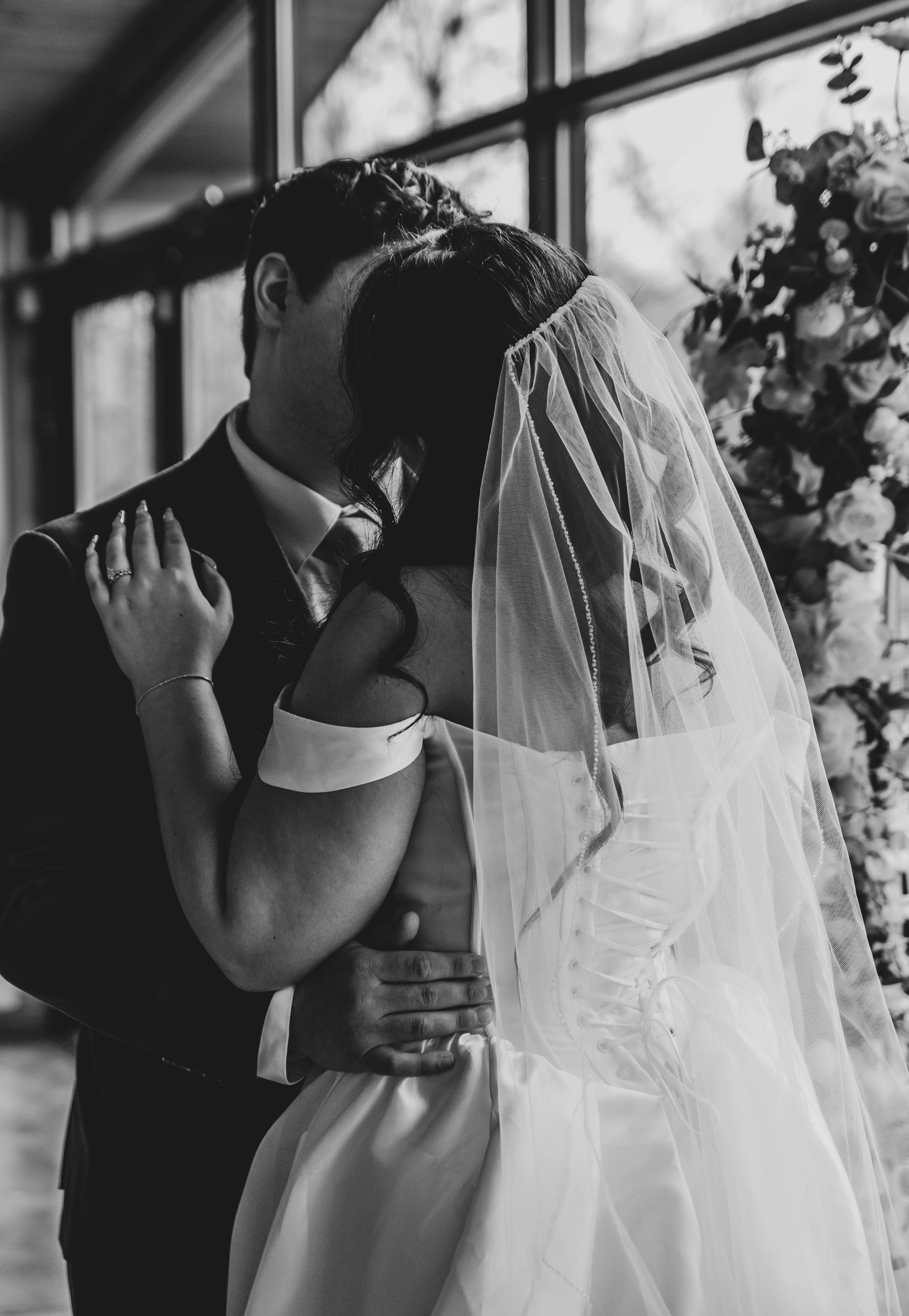 Black and white photo of a bride and groom sharing their first kiss at the altar, surrounded by floral arrangements.