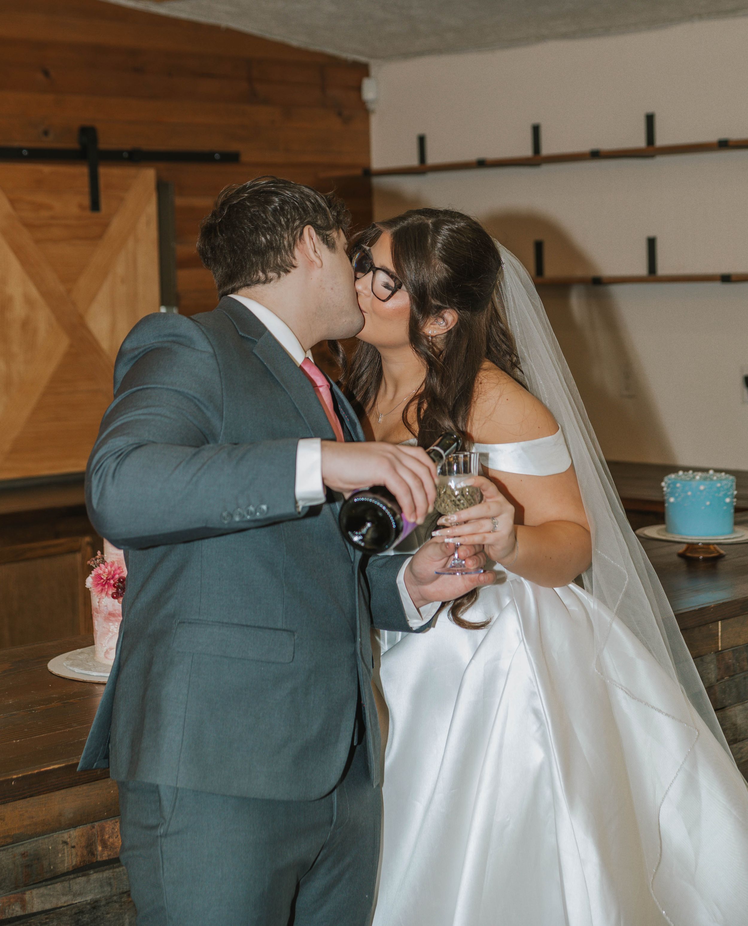 Newlywed couple sharing a kiss while pouring champagne, bride in white gown with veil, groom in gray suit.