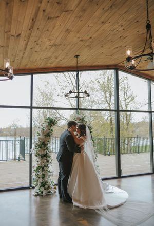 Bride and groom share first kiss at indoor wedding ceremony with floral arch and large windows overlooking scenic waterfront.
