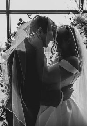 Black and white wedding photo of bride and groom sharing intimate moment under veil near floral arch.