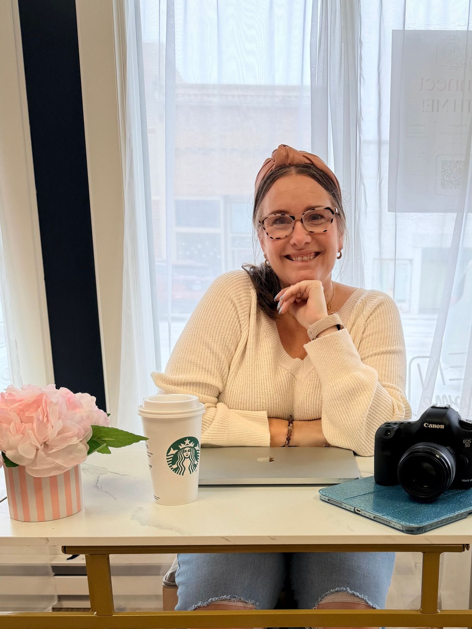 Woman smiling at desk with Starbucks cup, Canon camera, laptop, and pink flowers by bright window.