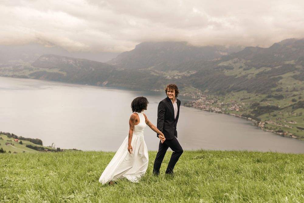 Bride and groom holding hands and walking together on a grassy hill overlooking a vast mountain lake in Lucerne.