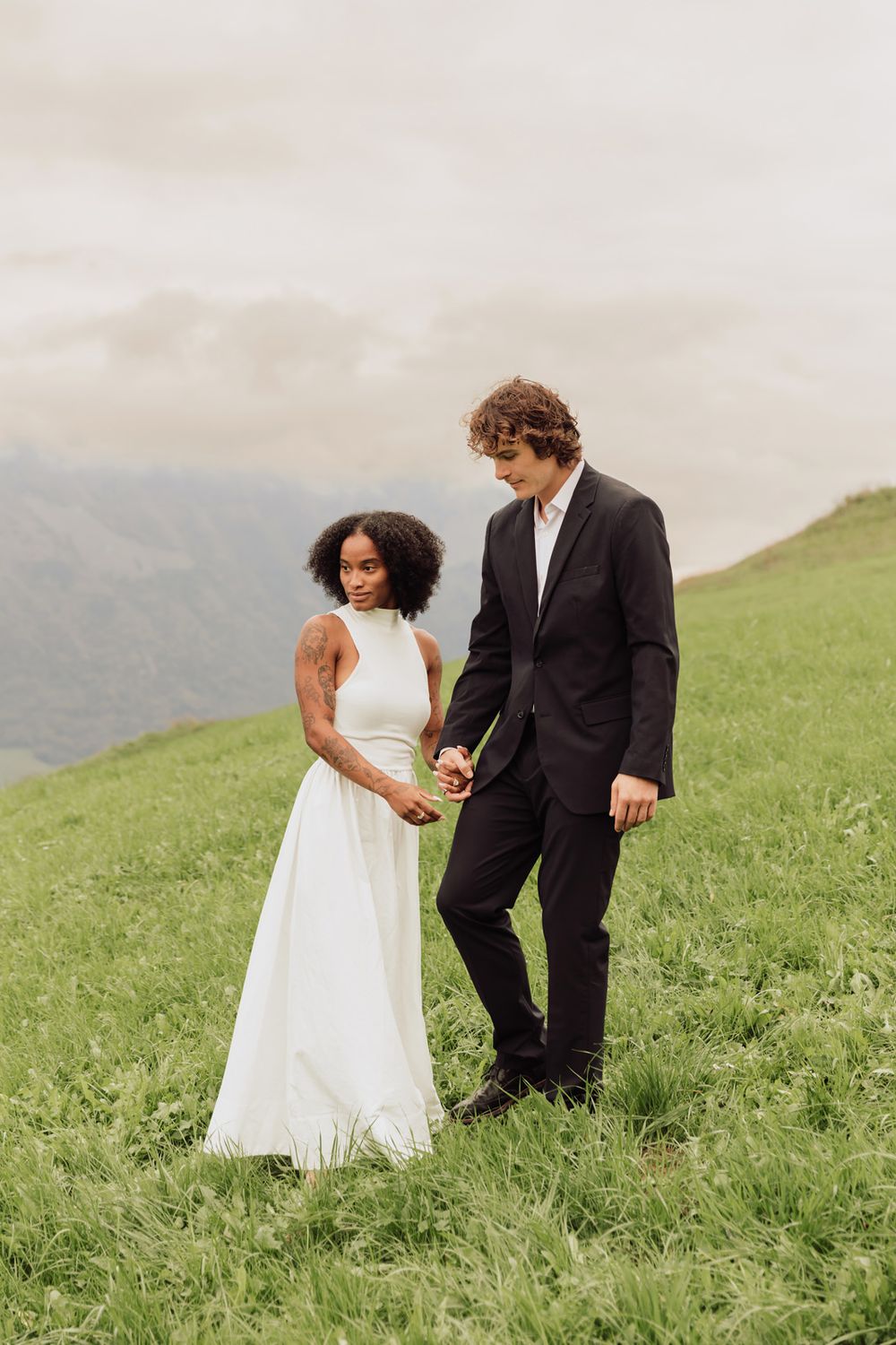Full-length vertical portrait of a bride and groom standing together on a lush green hillside.