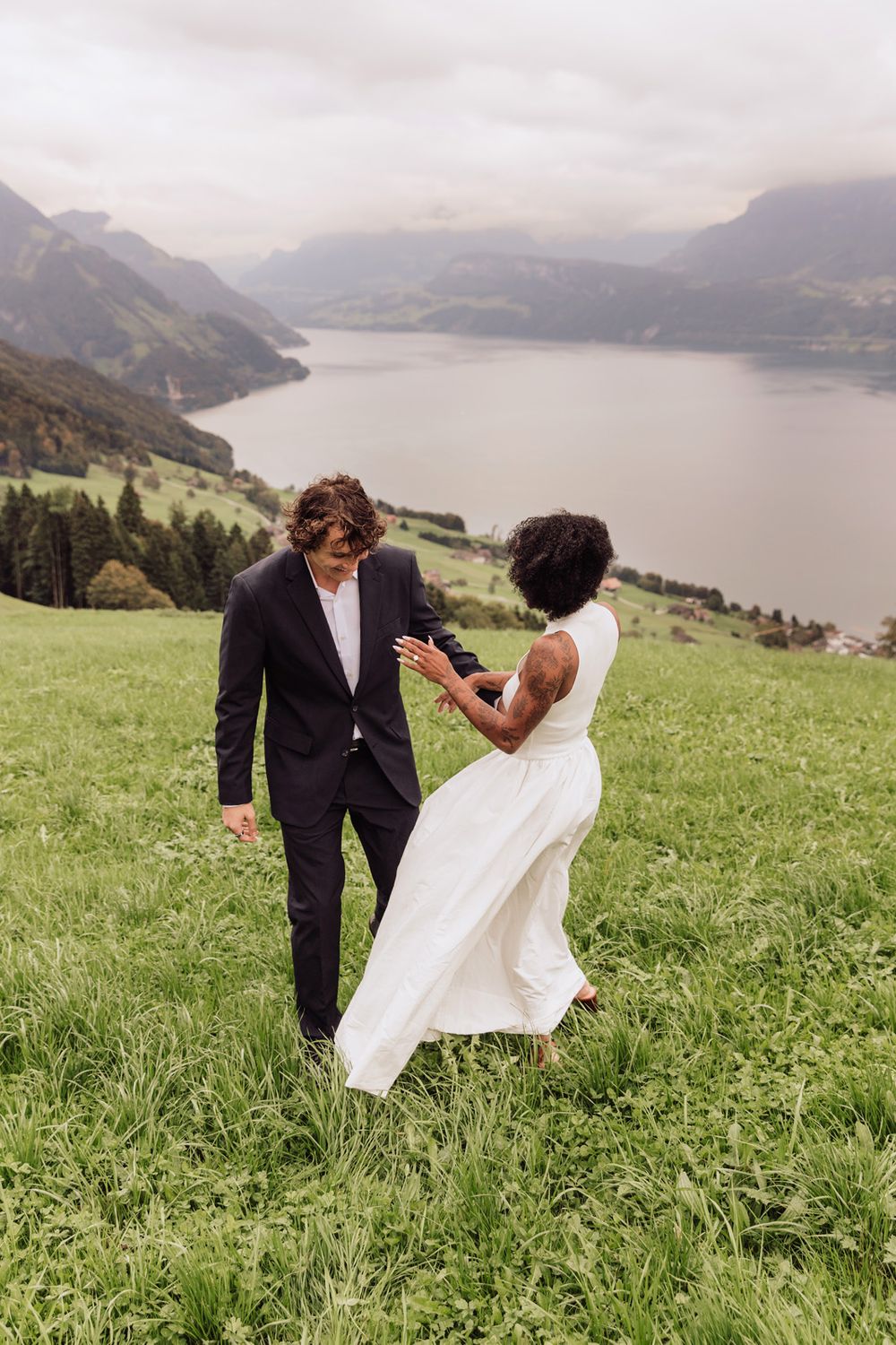 Wide landscape photo of a couple holding hands in a grassy field with a misty mountain lake background.