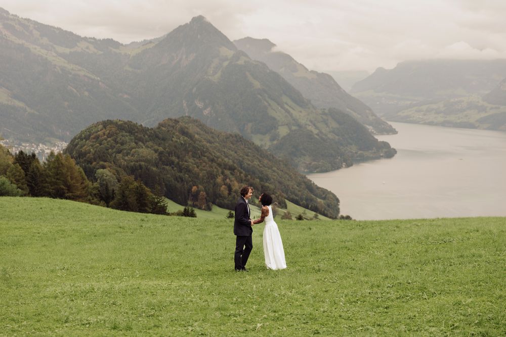 Cinematic landscape photo of a couple holding hands in a vast green field by a mountain lake.