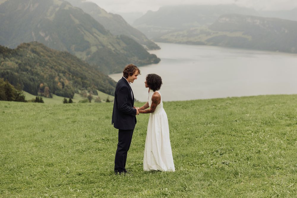 Wide shot of a couple standing together on a lush green hill with a sweeping lake and mountain backdrop.