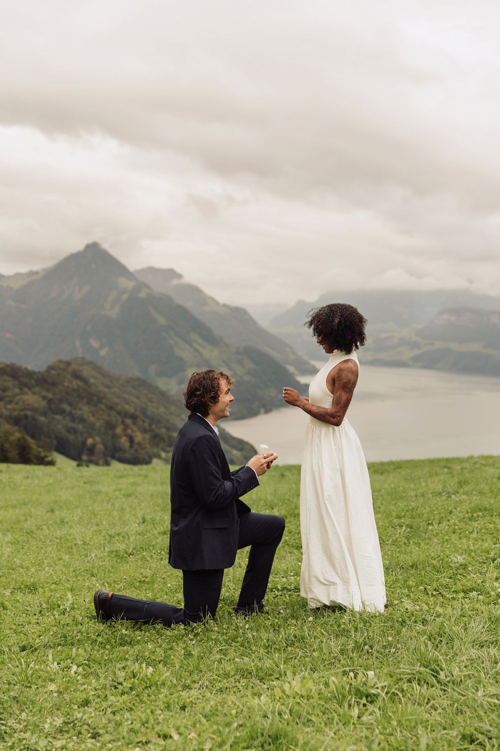 A man on one knee proposing to a woman on a grassy cliff overlooking a scenic mountain valley.