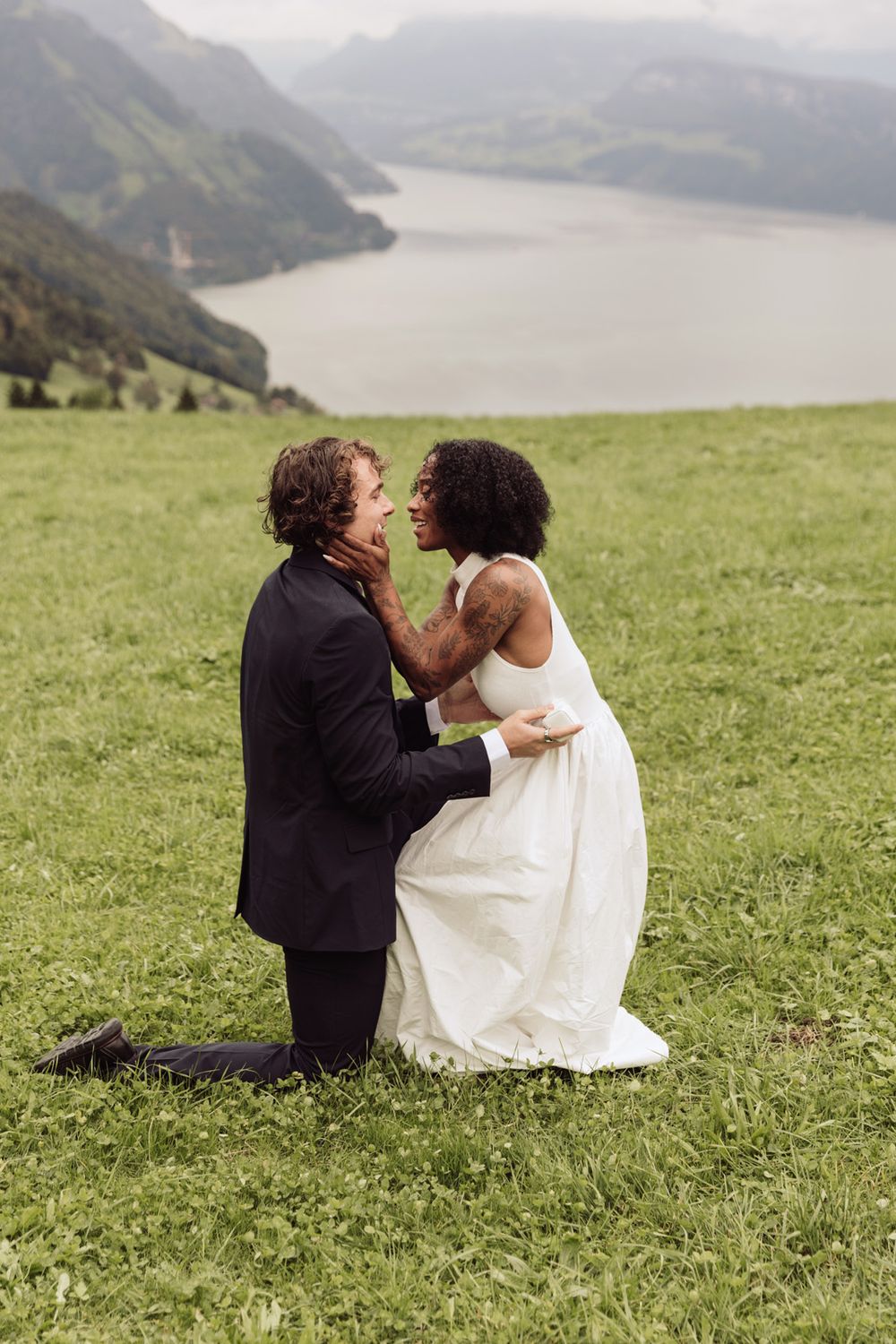 Newly engaged couple sharing a hug on a scenic cliffside with a mountain and water backdrop.
