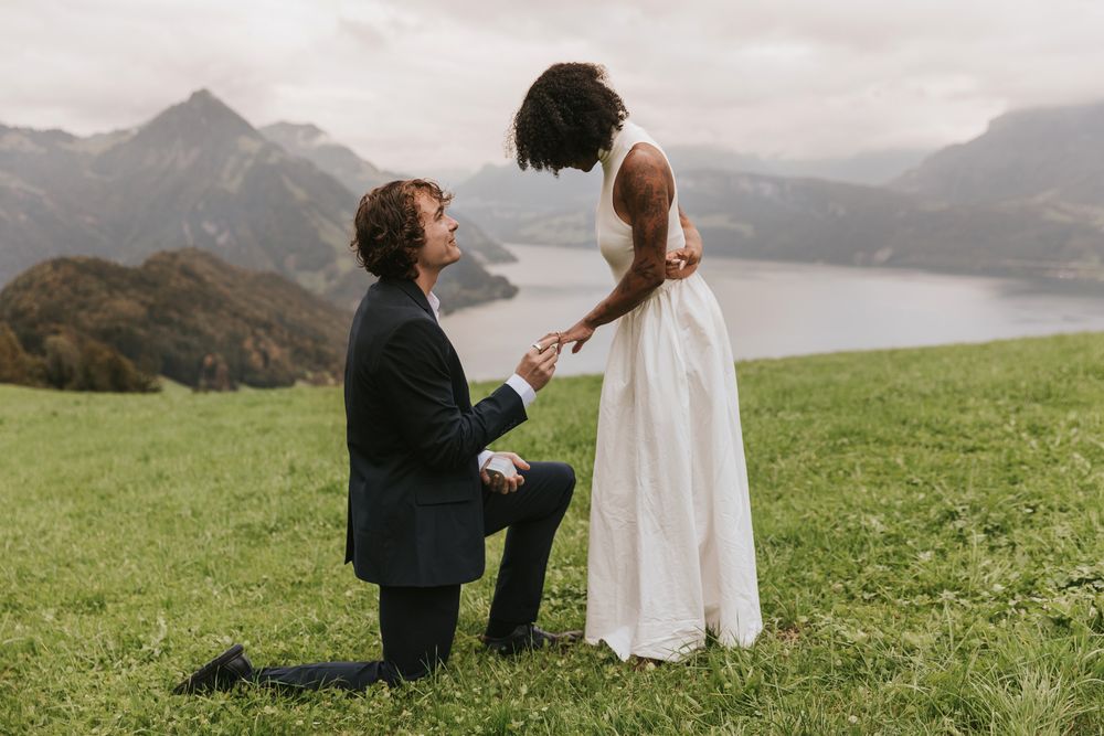 Wide shot of a man proposing to a woman on a grassy hilltop overlooking a scenic lake.