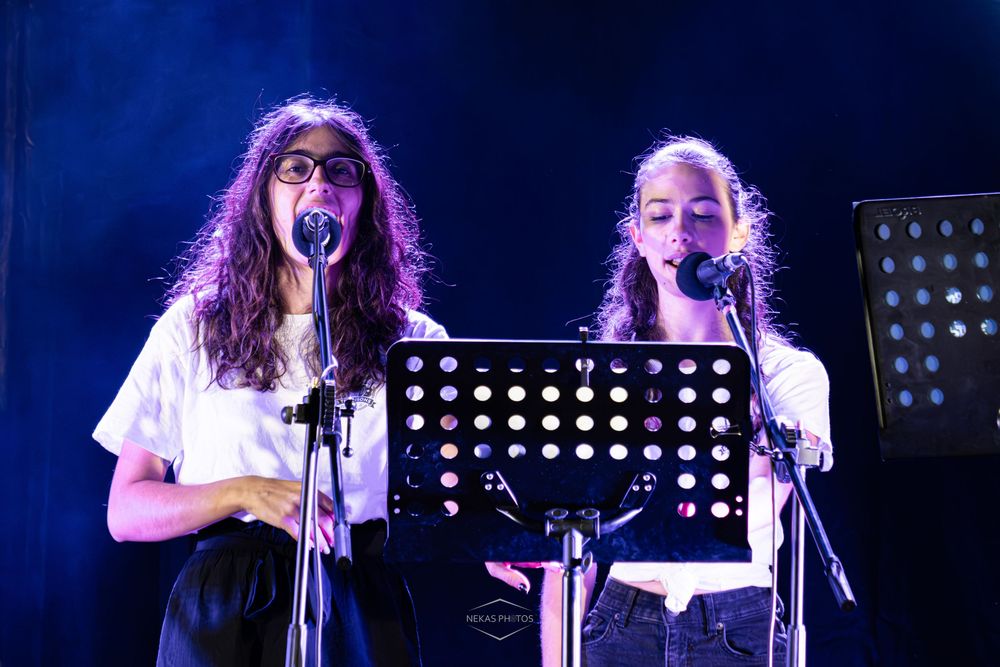 Two young women singing on stage with microphones and a music stand under blue stage lighting.