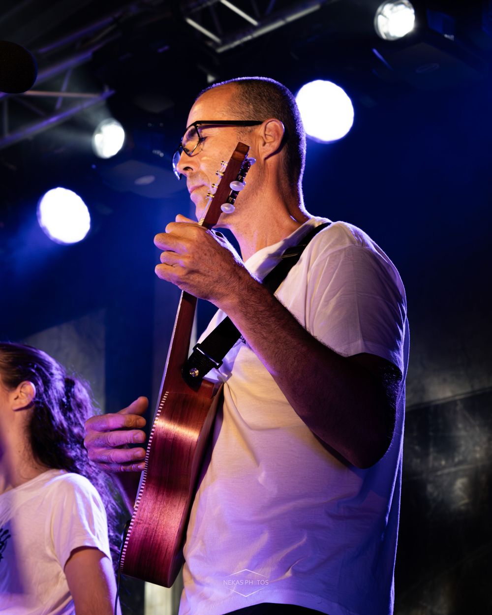 Man with glasses playing acoustic guitar on stage under bright lights wearing a white t-shirt.