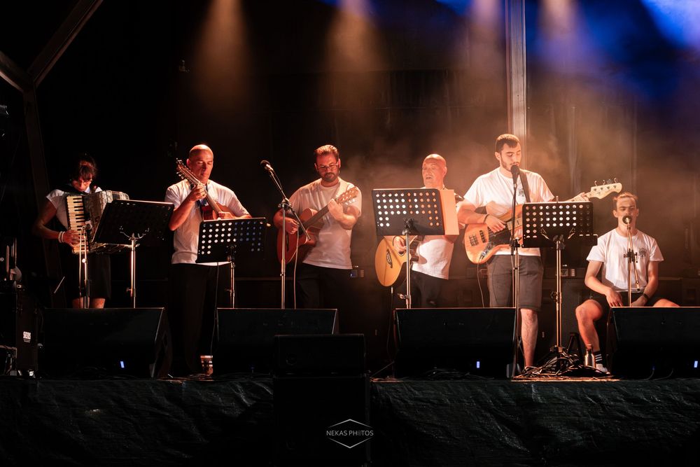 Live band performing on stage with guitars and brass instruments under colorful lights at an outdoor concert.