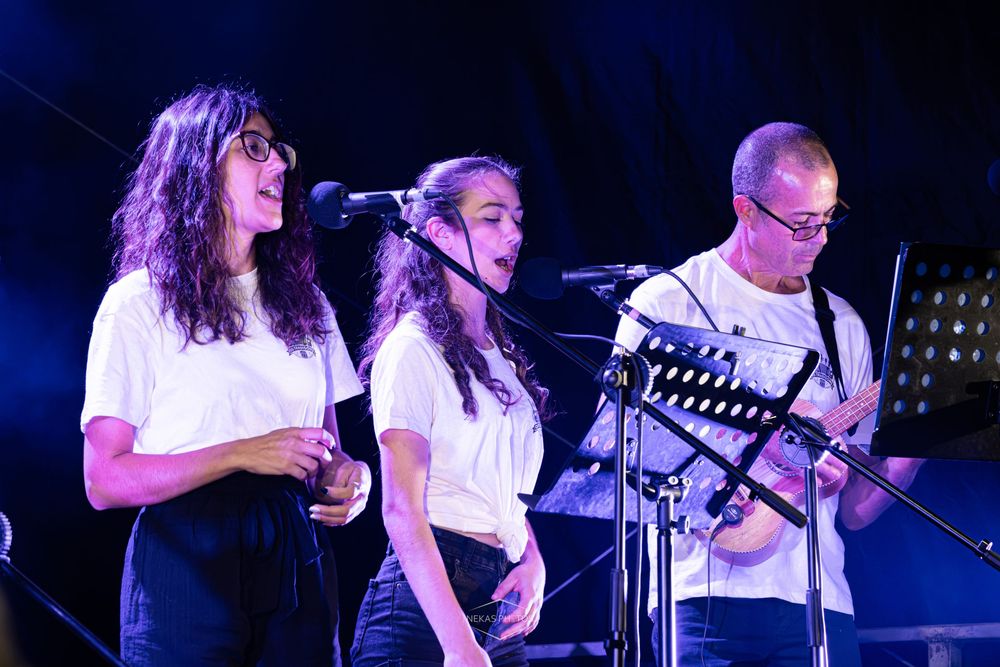 Three performers in white t-shirts singing on stage at an outdoor concert under blue lights.