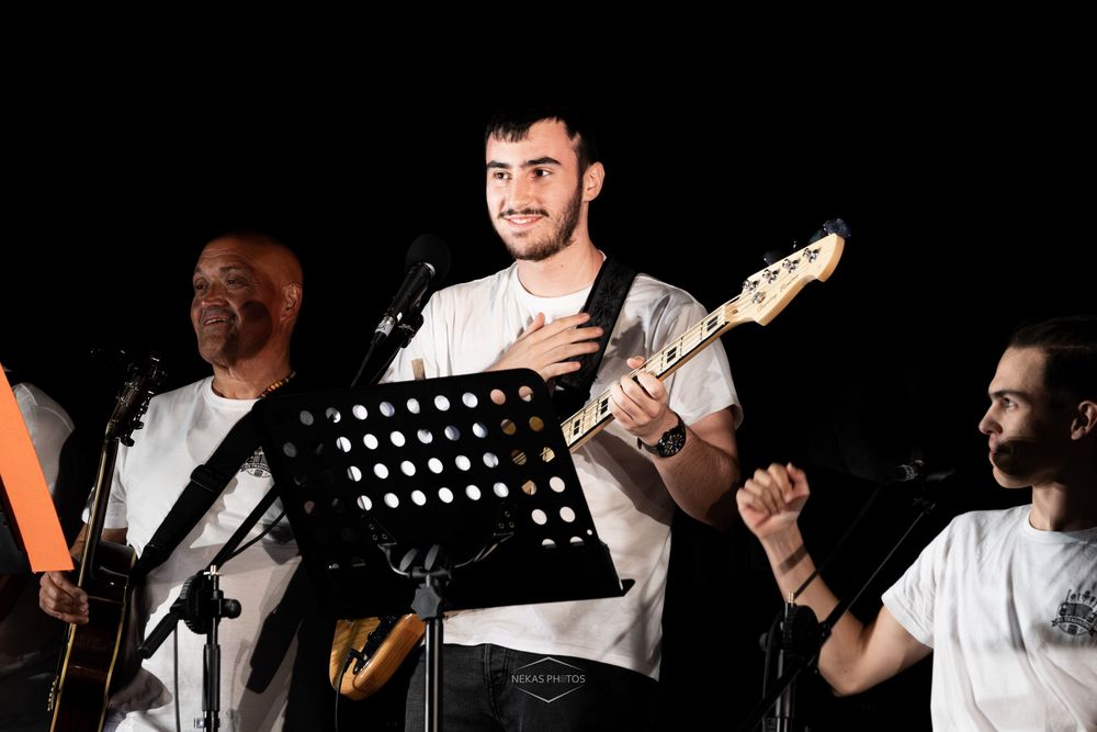 Three musicians performing on stage, center guitarist in white shirt smiling and holding electric guitar under bright stage lights.