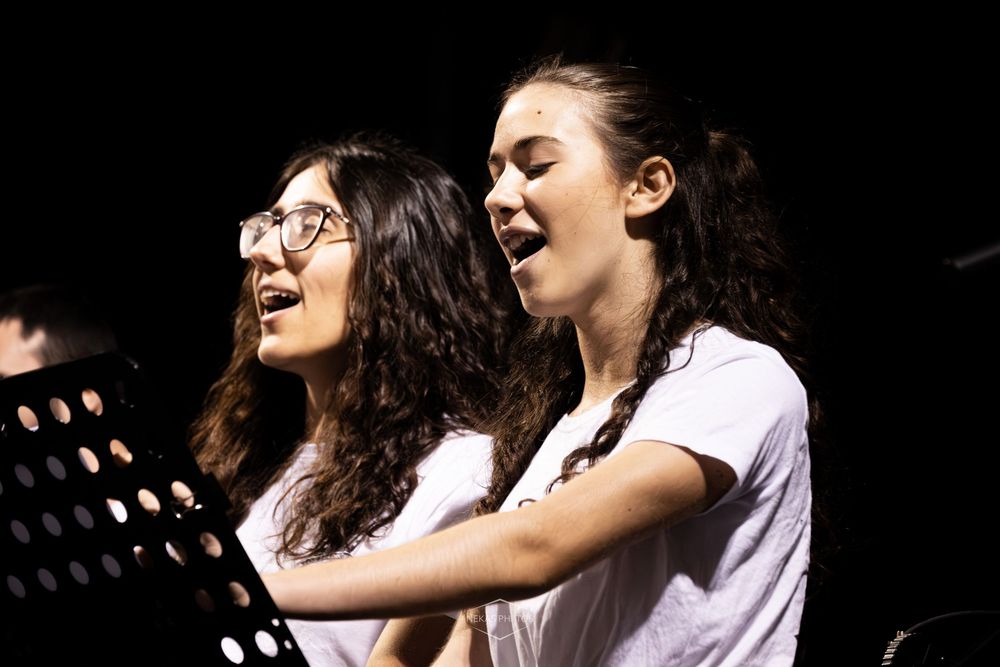Two young women singing together at a music stand performance, one wearing glasses, both with eyes closed.